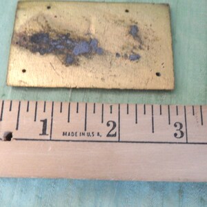 May include: A brass plaque with a rectangular shape and a slightly textured surface. The plaque is approximately 1 inch by 2 inches in size. The plaque has four holes in the corners and shows signs of wear and tear.