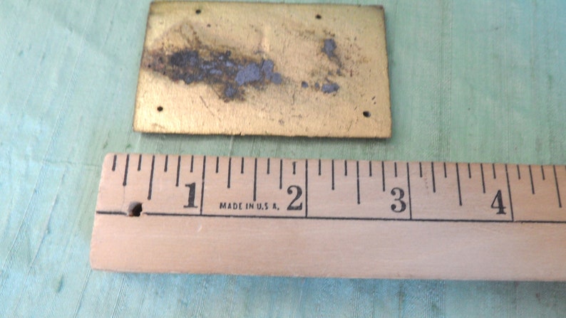 May include: A brass plaque with a rectangular shape and a slightly textured surface. The plaque is approximately 1 inch by 2 inches in size. The plaque has four holes in the corners and shows signs of wear and tear.