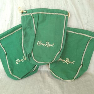 May include: Three green drawstring bags with white stitching and the words "Crown Royal" embroidered in a cursive font. The bags are made of a soft fabric and have a drawstring closure at the top.