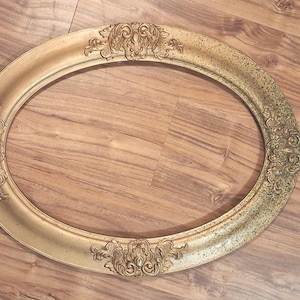 May include: An oval-shaped picture frame with a metallic gold finish. The frame features ornate, raised scrollwork and floral relief detailing at the top, bottom, and sides. The surface shows a gradient of color from smooth gold to a darker, speckled patina.