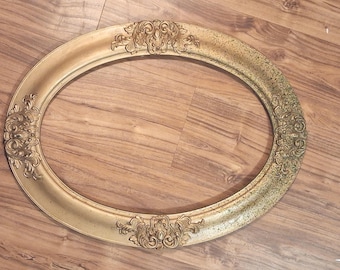 May include: An oval-shaped picture frame with a metallic gold finish. The frame features ornate, raised scrollwork and floral relief detailing at the top, bottom, and sides. The surface shows a gradient of color from smooth gold to a darker, speckled patina.