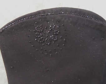Pode incluir: Close-up de um item de tecido preto, possivelmente uma manga ou punho, adornado com pequenas contas escuras. A borda é forrada com uma fileira de contas, e um design floral é criado com contas agrupadas.