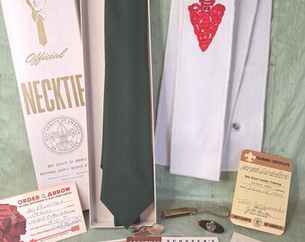 May include: A dark green necktie in a box labeled Official Necktie Boy Scouts of America, a white sash with a red embroidered arrow, a metal tie clip, and various vintage Boy Scouts of America training certificates and progress cards.
