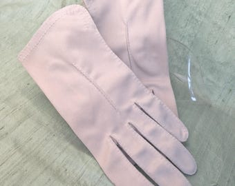 May include: A pair of light pink gloves. The gloves are made of a smooth fabric and have a classic design with individual finger slots. The gloves are likely for formal wear or a vintage aesthetic.