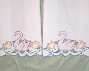 May include: Two white rectangular fabric panels featuring embroidered swan motifs at the bottom. Each swan is outlined in pink, flanked by yellow flowers and green lily pads, with a scalloped blue embroidered edge along the bottom hem.