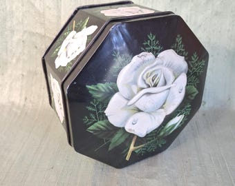 May include: An octagonal black metal tin featuring a large white rose illustration on the lid and sides. The floral design includes green leaves and delicate fern-like foliage. The container has a removable lid and a dark, glossy finish.