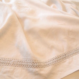 Puede incluir: Un mantel de lino blanco con un delicado borde de encaje. El mantel tiene un diseño bordado sutil en el centro.