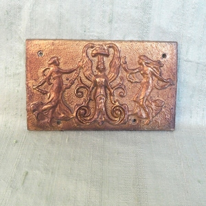 May include: A rectangular plaque with a raised design of three figures in a stylized dance. The figures are made of a copper-colored metal and are set against a gold background.