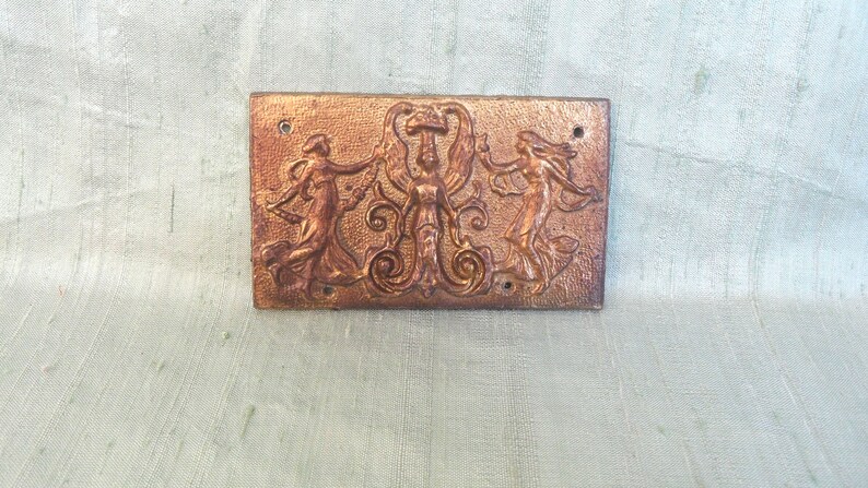 May include: A rectangular plaque with a raised design of three figures in a stylized dance. The figures are made of a copper-colored metal and are set against a gold background.