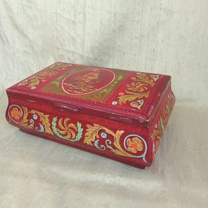 May include: A rectangular red tin box with ornate gold and green floral and scrollwork designs. A central oval features a sailing ship. The box has a curved base and lid, and is likely vintage or antique.