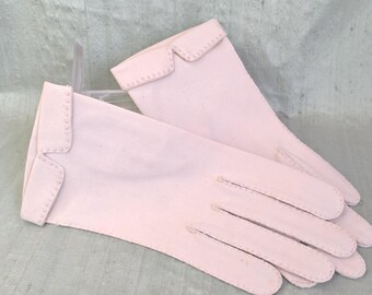 May include: A pair of pale pink gloves. The gloves have a classic design with a scalloped edge on the cuff, adorned with small, evenly spaced decorative details. The gloves are made of a soft, smooth material.