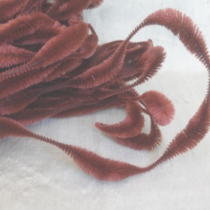 May include: Close-up of burgundy-colored craft stems, possibly chenille stems or pipe cleaners, arranged on a light-colored surface. The stems have a fuzzy texture and are curved and looped, suggesting a decorative or crafting purpose.