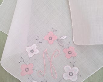 May include: A white square handkerchief featuring a central embroidered monogram letter N in pink thread. The letter is surrounded by a circular wreath of embroidered flowers in shades of pink and white, with gray stems and leaves.