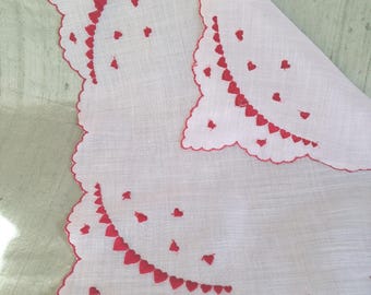 May include: A white fabric square with scalloped edges trimmed in red thread. The textile features embroidered red heart motifs arranged in curved lines and scattered patterns across the surface, with one corner folded over to reveal the design.