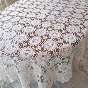 May include: An oval white tablecloth featuring an intricate crochet pattern composed of repeating circular motifs connected by a delicate openwork mesh design. The lace fabric drapes over the edges of a wooden table, showcasing a detailed texture.