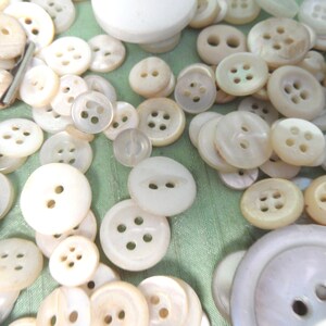 May include: A large collection of white pearl buttons of various sizes and styles. The buttons are arranged in a random pattern on a green surface.