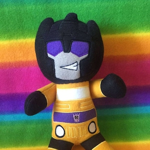 Könnte beinhalten: Ein gelbes und schwarzes Plüschtier eines Roboters mit einem violetten Gesicht und einem violetten Symbol auf der Brust. Das Spielzeug sitzt auf einer Regenbogen-Decke.