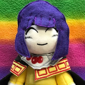 May include: A plush toy of a character with purple hair, a yellow shirt, black trousers, and a black cape. The toy is smiling and has red accents on its shirt.