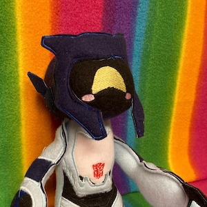 Könnte beinhalten: Ein Plüschtier eines Robotercharakters mit einem blauen Helm, einem weißen Körper und schwarzen Akzenten. Der Roboter hat ein gelbes Gesicht und ein rotes Autobot-Symbol auf der Brust.