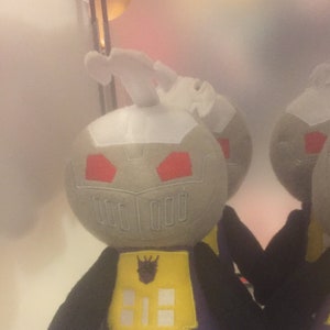 Puede incluir: Dos peluches de un personaje robot con cabeza gris, ojos rojos, pecho amarillo con un símbolo negro y piernas moradas y negras.