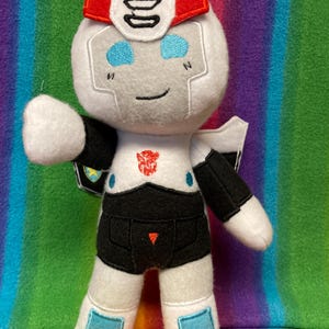 Op de afbeelding: Een pluche robotknuffel met een witte kop, rode accenten, blauwe ogen en een zwart-wit lichaam. Op de borst staat een rood Autobot-symbool. De armen en benen zijn wit met zwarte en blauwe details en de figuur heeft een kleine zwarte glimlach.