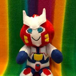 May include: A plush toy robot with a red head, white body, and blue accents. The robot has a smiling face with blue eyes and yellow details. The toy is sitting on a rainbow striped background.