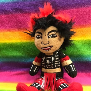 May include: A plush toy doll with black hair, red and black clothing, and a black and white skeleton design. The doll is sitting on a rainbow striped blanket.