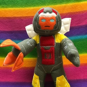 Może przedstawiać: Pluszowa zabawka robot w kolorze szarym i żółtym z czerwonymi akcentami. Robot ma biały hełm z czerwoną twarzą i niebieskimi oczami. Robot ma skrzydła i pazur na każdym ramieniu.