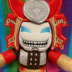 Könnte beinhalten: Ein Plüschtier, das eine Figur mit einem rot-braunen Samurai-Helm mit silbernem, kreisförmigem Wappen darstellt. Die Figur hat ein hellgraues Gesicht, leuchtend blaue gestickte Augen und ein breites, zahniges Grinsen in rot-brauner Rüstung.