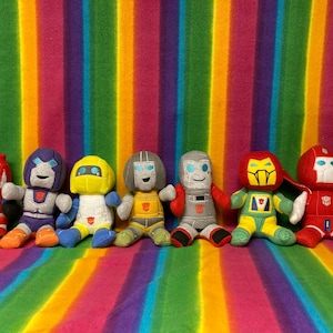 May include: Seven plush toy figures in various colors, including red, blue, yellow, and gray, are arranged in a row. Each figure has a unique design, with robot-like features and distinct facial expressions. The background is a vibrant rainbow-striped fabric.