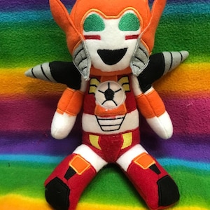 May include: A plush toy of a cartoon robot with orange, red and white colouring. The robot has a large, toothy grin and green eyes. The robot is sitting on a rainbow striped blanket.