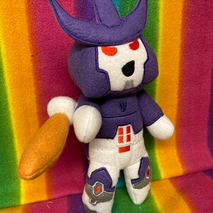 Könnte beinhalten: Ein handgefertigtes Plüschtier in Form eines Robotercharakters. Das Spielzeug hat einen lila Kopf mit Krone, rote Augen und ein weißes Gesicht. Es trägt ein lila-weißes Outfit mit orangefarbenen und grauen Akzenten und hält eine orangefarbene Waffe.