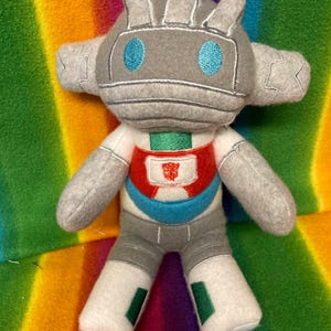 Könnte beinhalten: Ein handgefertigtes Plüschtier-Roboter in Grau, Weiß, Rot und Blau. Der Roboter hat einen grauen Kopf mit blauen Augen, graue Arme und weiße Beine mit grünen Akzenten. Die Brust des Roboters zeigt ein rotes Emblem. Das Spielzeug befindet sich auf einem Regenbogenhintergrund.