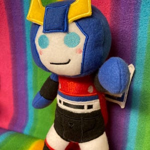 Puede incluir: Un juguete de peluche azul y rojo con cuernos amarillos, ojos blancos y una cara sonriente. El juguete lleva una falda negra y botas rojas.