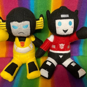 Könnte beinhalten: Zwei Transformers-Plüschfiguren. Eine ist gelb und schwarz, die andere rot und schwarz. Beide haben detaillierte Gesichtszüge und das Autobot-Symbol. Sie sind vor einem bunten, regenbogenfarbenen Hintergrund platziert.