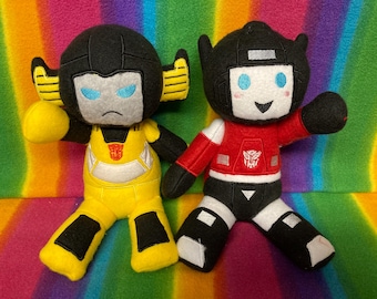 Puede incluir: Dos peluches de Transformers. Uno es amarillo y negro, el otro rojo y negro. Ambos tienen rasgos faciales detallados y el símbolo Autobot. Se colocan sobre un fondo colorido con rayas de arcoíris.