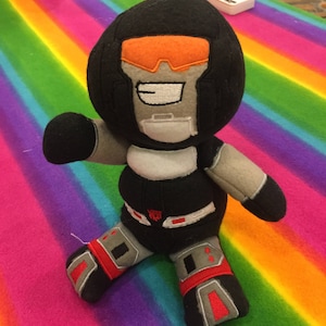 Könnte beinhalten: Schwarzes Plüschtier eines Roboters mit orangefarbenem Visier und einem lächelnden Gesicht. Das Spielzeug sitzt auf einer regenbogenfarbenen gestreiften Oberfläche. Der Roboter hat einen schwarzen und grauen Körper mit roten Akzenten und ein silbernes Emblem auf seiner Brust, auf dem "Transformers" steht.