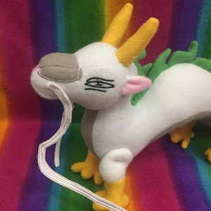 May include: White plush dragon with yellow horns, green scales, and a long white tail. The dragon has a gray snout and pink ears.