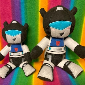 Könnte beinhalten: Zwei Plüschtiere eines Robotercharakters, eines größer als das andere. Beide sind schwarz-weiß mit blauen Akzenten und haben ein lächelndes Gesicht. Das größere Spielzeug ist etwa 30 cm groß und das kleinere etwa 15 cm.