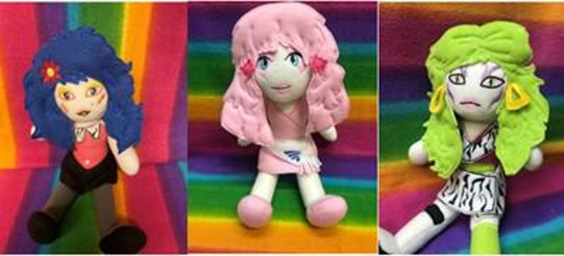 May include: Three handmade plush dolls with anime-inspired features. The dolls have colorful hair, detailed facial features, and unique outfits. The background is a vibrant rainbow pattern, enhancing the dolls' colorful appearance.