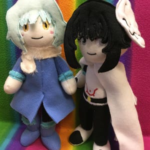 May include: Two handmade plush dolls. One doll is wearing a blue coat, a white scarf, and a light blue hat. The other doll is wearing a black outfit with a white cape and a white headpiece. Both dolls have embroidered faces.