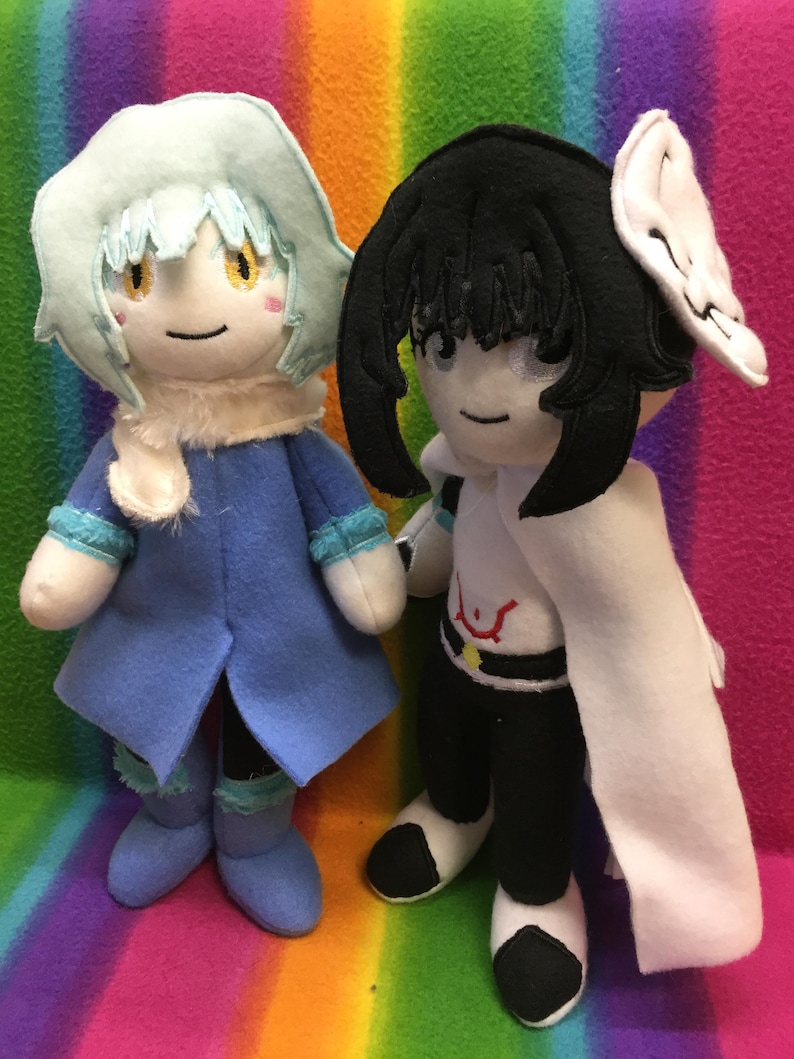 May include: Two handmade plush dolls. One doll is wearing a blue coat, a white scarf, and a light blue hat. The other doll is wearing a black outfit with a white cape and a white headpiece. Both dolls have embroidered faces.