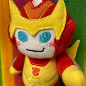 May include: A plush toy of a character with a red and yellow color scheme, featuring a helmet-like headpiece with yellow accents. The toy has blue eyes, a white face, and a small Autobot symbol on the chest. The background is a rainbow of colors.