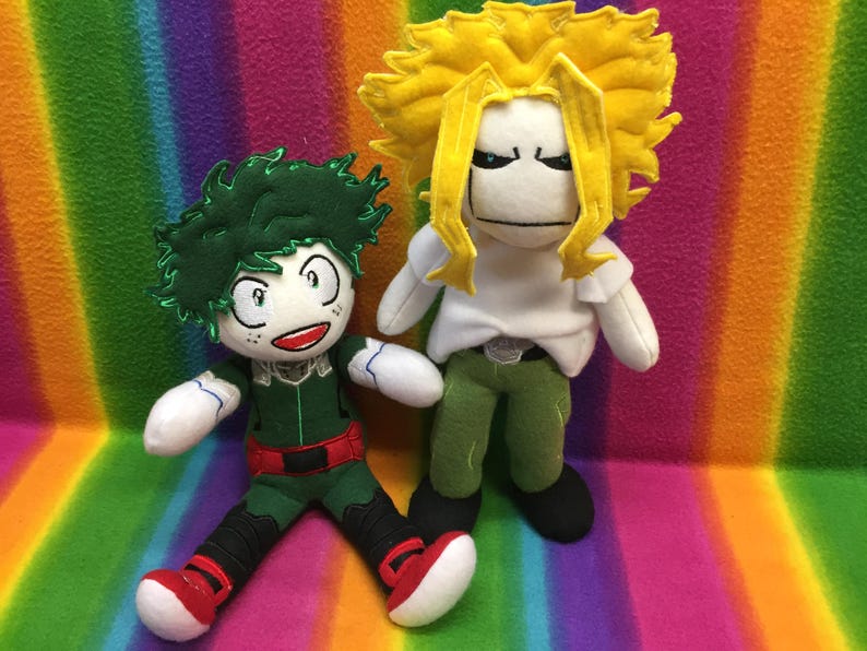 May include: Two plush dolls of anime characters. One doll has green hair, a green and red outfit, and red shoes. The other doll has yellow hair, a white shirt, green pants, and black shoes. The dolls are on a rainbow-colored background.
