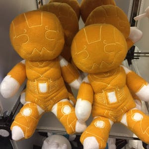 Könnte beinhalten: Eine Gruppe von fünf Plüschtieren in Form von Robotern. Die Spielzeuge sind gelb mit weißen Akzenten und haben ein einfaches, cartoonartiges Design.