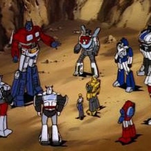 May include: A group of eight cartoon robots, including Optimus Prime, in various poses. The robots are all different colors and have different designs. They are standing on a brown, rocky surface.