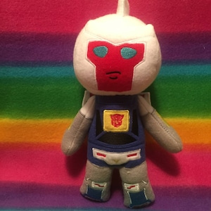 Puede incluir: Un juguete de peluche de un robot con cabeza blanca, cara roja, cuerpo azul y brazos y piernas grises. El robot tiene un símbolo Autobot amarillo en el pecho.