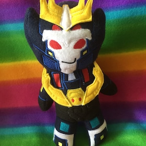 Könnte beinhalten: Ein Plüschtier-Roboter mit schwarzem Körper, weißem Gesicht und roten Augen. Der Roboter hat eine gelbe Krone und Akzente und steht auf einer regenbogenfarbenen Oberfläche. Das Spielzeug ist aus weichem Stoff gefertigt und hat gestickte Details.