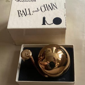 Peut inclure: Un ancien poudrier Wadsworth Ball and Chain dans sa boîte d'origine. Le poudrier est de couleur or brillant, sphérique, et attaché à une petite chaîne. La boîte est blanche avec du texte noir et un dessin d'une boule et d'une chaîne.