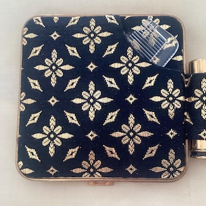 May include: Vintage vanity set with a black and gold floral pattern. The set includes a compact, a lipstick holder, and a comb. The compact is square with a gold-toned frame. The lipstick holder is cylindrical, and the comb is clear plastic.